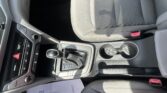 Interior of a car showing the center console with gear shifter, cup holders, and a gray fabric seat; sunlight casts shadows on the seat edge — passenger-side area visible.