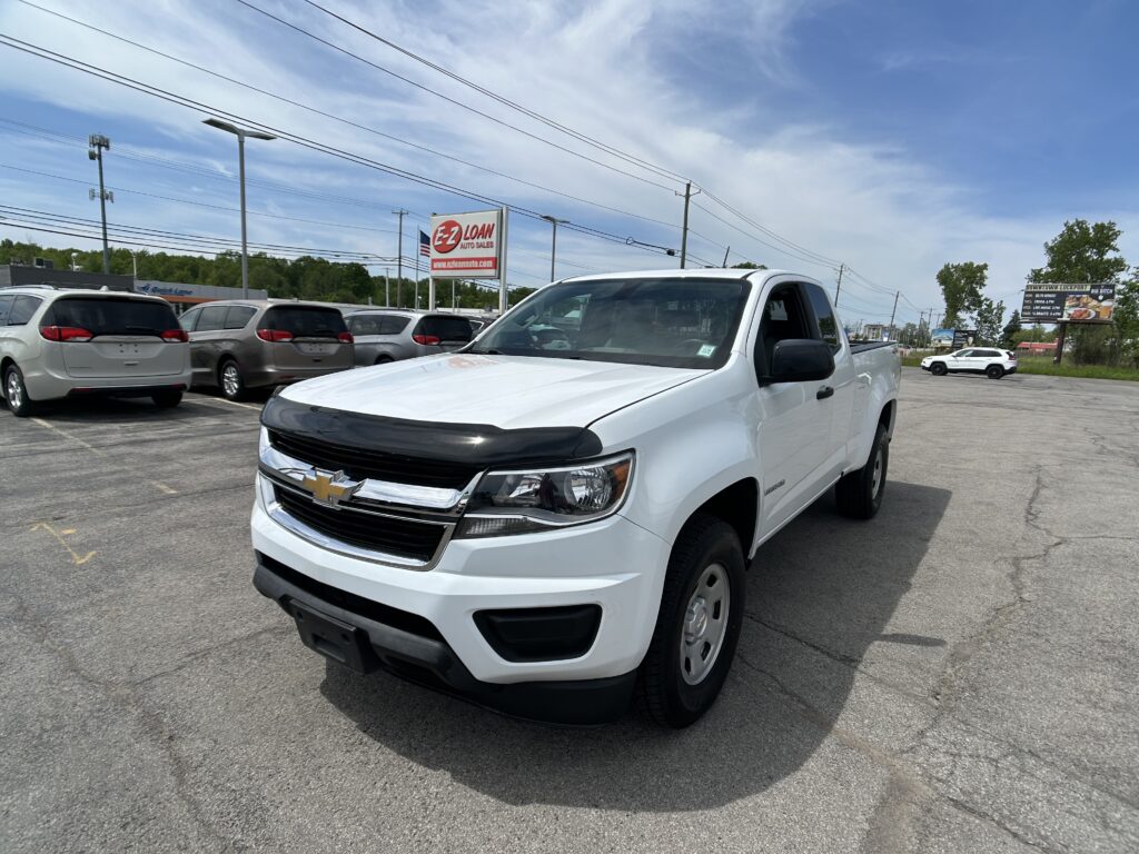 White Chevrolet pickup at a dealership lot with multiple minivans and a big sign in the background, under a blue sky with power lines above.