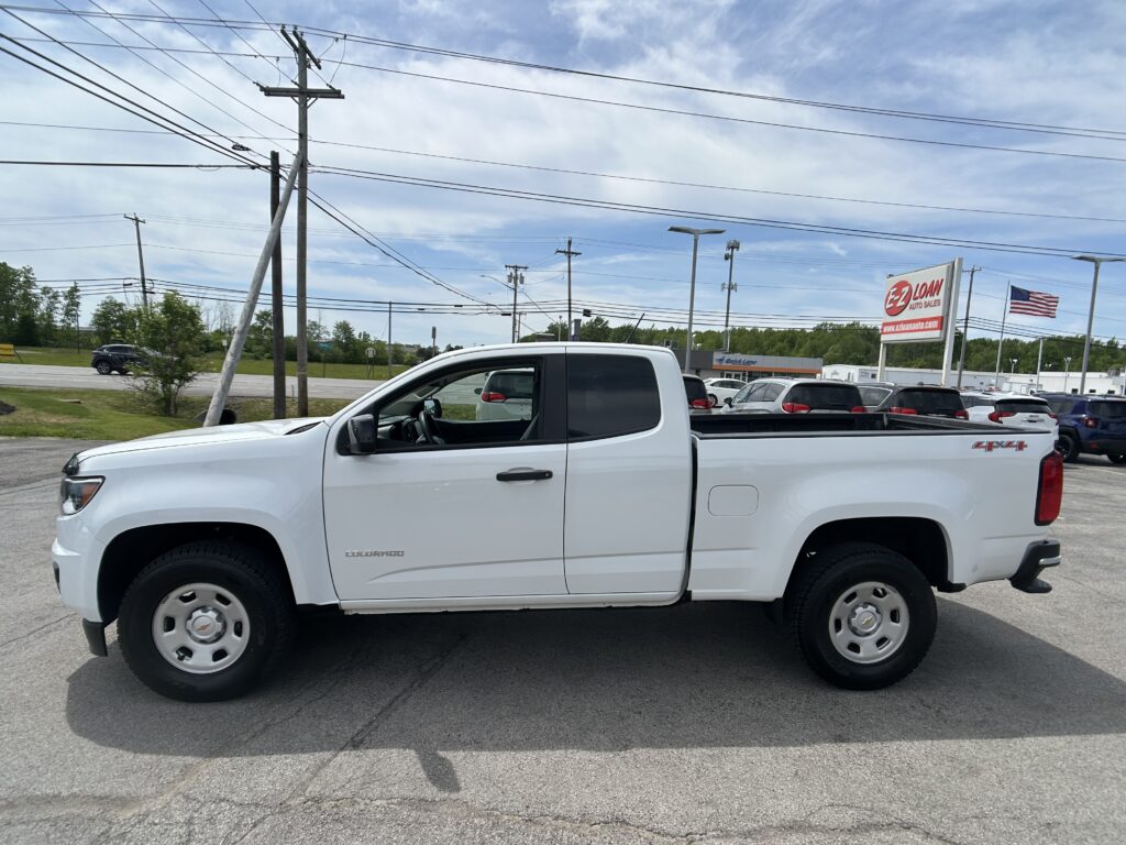 White Chevrolet Colorado pickup in a parking lot, with power lines overhead and an E-Loan Auto Sales sign in the background and an American flag nearby.