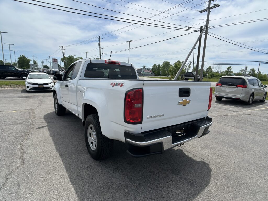 Rear three-quarter view of a white Chevrolet Colorado pickup in a car dealership lot, with power poles and wires overhead.