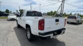 Rear three-quarter view of a white Chevrolet Colorado pickup in a car dealership lot, with power poles and wires overhead.