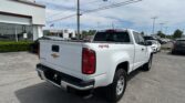 Rear of a white Chevrolet pickup truck in a dealership lot, with a Toyota dealership building in the background and an American flag overhead.
