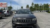 Front view of a black Jeep SUV in a dealership parking lot with other cars and a blue sky above.