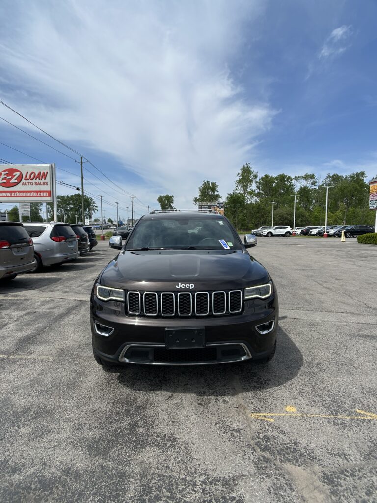 Front view of a black Jeep SUV in a dealership parking lot with other cars and a blue sky above.