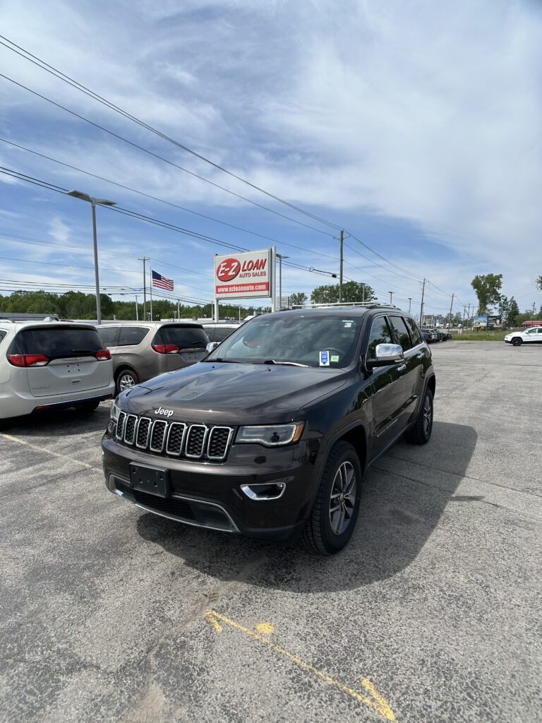 Dark Jeep SUV in the foreground at a car dealership, with an EZ Loan Auto Sales sign and an American flag in the background.