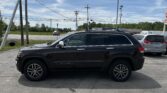 Side view of a black Jeep Grand Cherokee SUV parked in a parking lot with power lines overhead and a blue sky Behind it, other cars visible in the background.