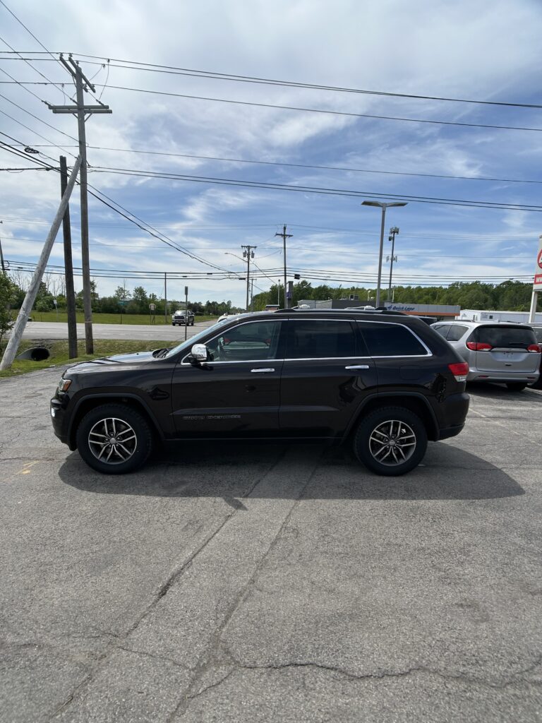 Side view of a black Jeep Grand Cherokee SUV parked in a parking lot with power lines overhead and a blue sky Behind it, other cars visible in the background.