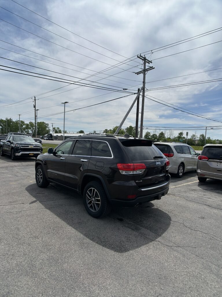 Black Jeep Grand Cherokee in a parking lot with other cars and tall utility poles overhead against a blue sky.