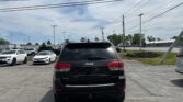 Rear view of a black Jeep SUV in a dealership parking lot with other cars and a blue sky above, power lines crisscrossing the scene.