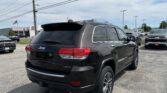 Black Jeep Grand Cherokee Limited SUV parked in a dealership lot with power lines overhead and a building in the background.