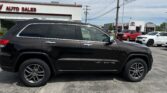 Black Jeep Grand Cherokee SUV parked in a dealership lot with a white auto sales building and other cars in the background under a blue sky.