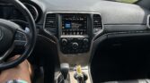 View from the driver’s seat of a car: dashboard, steering wheel, and center touchscreen visible with a parking lot of cars outside the windshield.