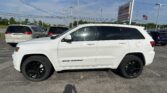 White Jeep Grand Cherokee in a dealership lot with other vehicles and an EZ Loan Auto Sales sign in the background; American flag nearby.