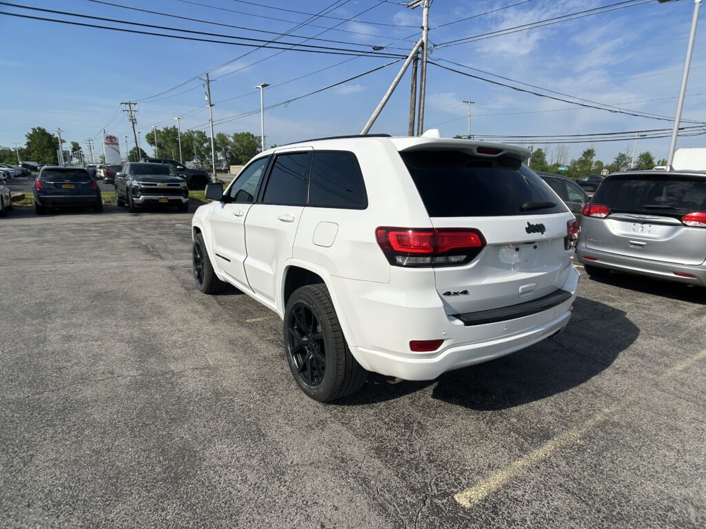 Rear three-quarter view of a white Jeep SUV in a car dealership lot with other vehicles and a Toyota sign in the background.