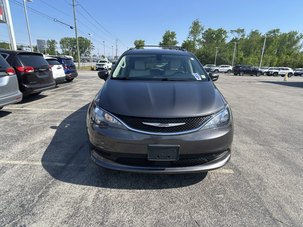 Front view of a gray Chrysler SUV with a roof rack in a crowded dealership lot under a clear blue sky, other cars parked nearby.