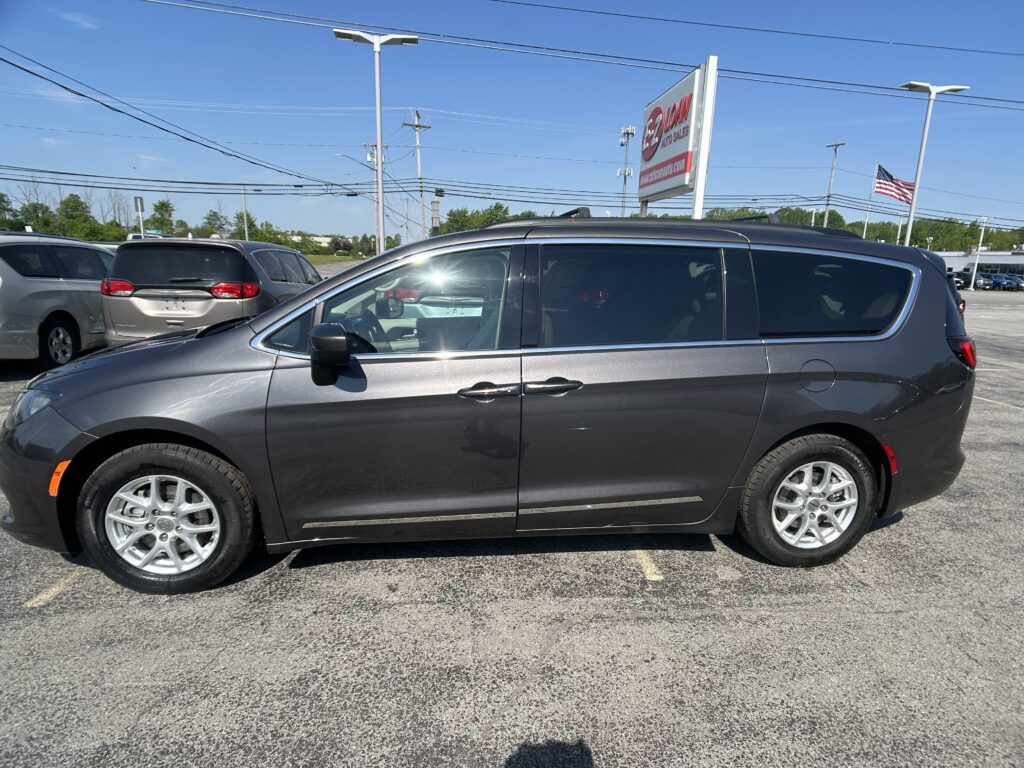 Side view of a dark gray SUV in a car dealership lot, with other vehicles, a tall sign, and an American flag in the background.