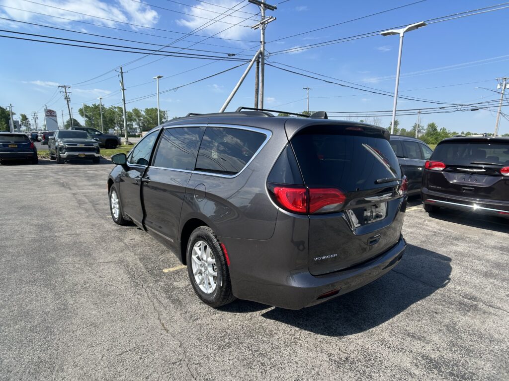 Rear three-quarter view of a gray Chrysler Voyager minivan in a dealership lot, with other cars and a Toyota sign in the background.