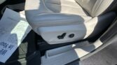 Beige leather car seat with perforated panels and electric adjustment controls; shadow across the seat and a paper floor mat nearby on the passenger side area.