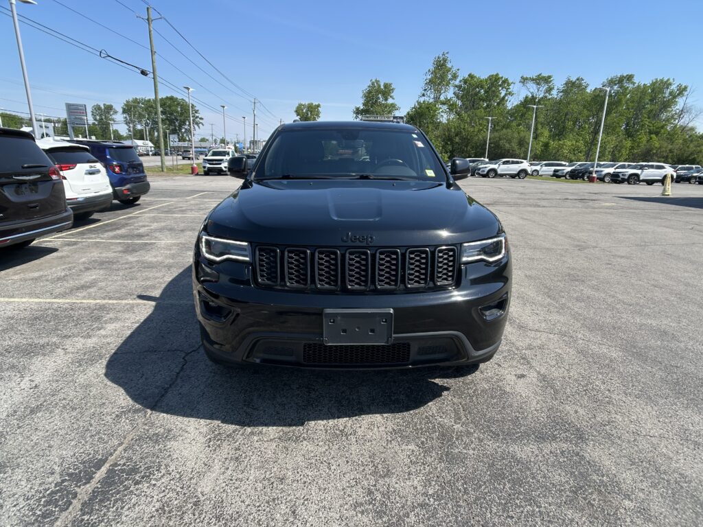 Front view of a dark Jeep SUV in a crowded car dealership lot on a sunny day, with other cars and trees in the background.