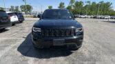 Front view of a dark Jeep SUV in a crowded car dealership lot on a sunny day, with other cars and trees in the background.