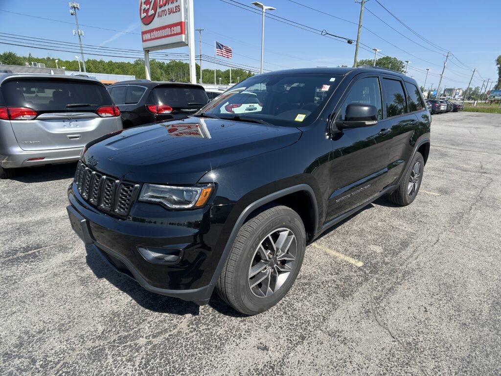 Front-right view of a black Jeep Grand Cherokee in a car dealership lot with other SUVs and a bright blue sky.