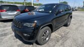 Front-right view of a black Jeep Grand Cherokee in a car dealership lot with other SUVs and a bright blue sky.