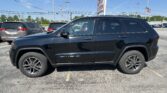 Black Jeep Grand Cherokee in a dealership parking lot with other cars, a large sign, and an American flag in a clear blue sky in the background.]