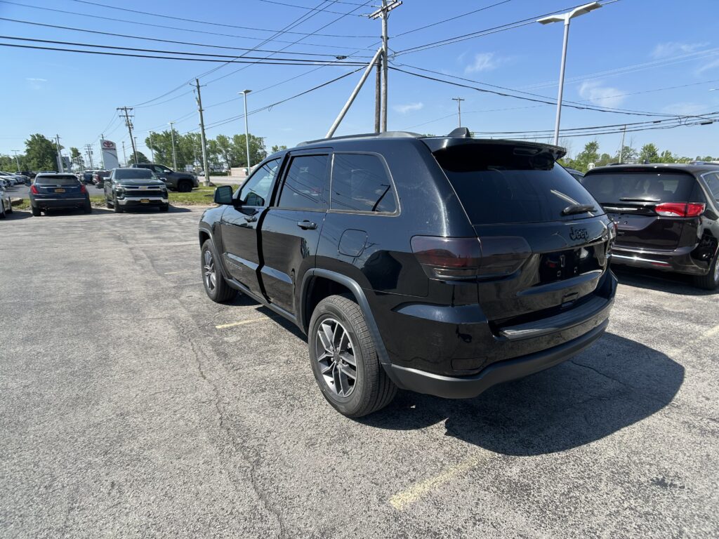 Black Jeep SUV parked in a crowded dealership lot under a clear blue sky with overhead power lines visible. The rear and side of the vehicle are prominent along with other cars in the background.