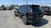 Black Jeep SUV parked in a crowded dealership lot under a clear blue sky with overhead power lines visible. The rear and side of the vehicle are prominent along with other cars in the background.