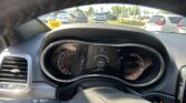 Driver's-seat view of the car’s dashboard with the speedometer and gauges, overlooking a parking lot with several cars outside. This shows the interior controls and the outside vehicles.