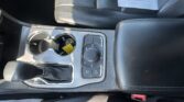 Center console of a car with two cup holders, a circular drive-mode knob, and a nearby gear selector; a paper document rests on the front passenger seat nearby.