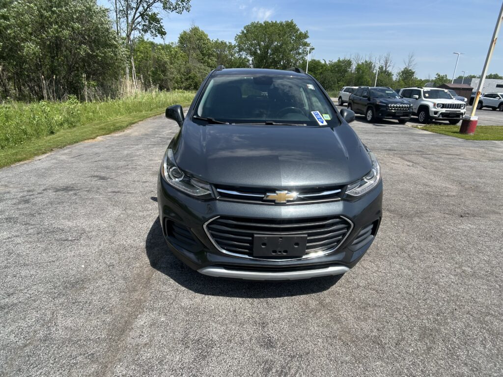 Front view of a dark gray Chevrolet SUV parked in a lot, with other cars and green trees in the background under a blue sky.