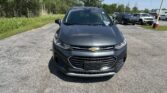 Front view of a dark gray Chevrolet SUV parked in a lot, with other cars and green trees in the background under a blue sky.