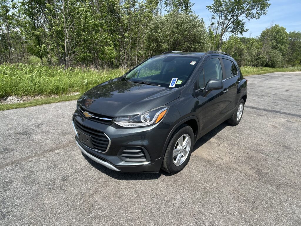 Dark gray Chevrolet SUV parked on a paved road beside a grassy, wooded area with green trees in the background.