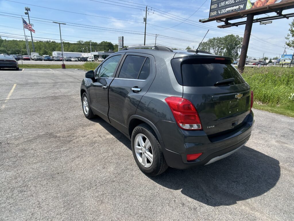 Rear three-quarter view of a gray Chevrolet Trax parked in a lot with a blue sky, flagpole, and billboards in the background.