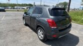 Rear three-quarter view of a gray Chevrolet Trax parked in a lot with a blue sky, flagpole, and billboards in the background.