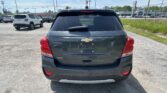 Rear view of a dark gray Chevrolet Trax LT parked in a lot, with other cars and a dealership in the background on a sunny day
