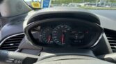 Driver's-eye view of the car dashboard with speedometer and tachometer, central display showing 0 mph, outside a parking lot through the windshield.