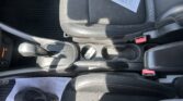 Interior of a car showing a black leather/posterior seat with stitching, center console with gear selector and cup holders, and scattered papers on the seat and floor mats.