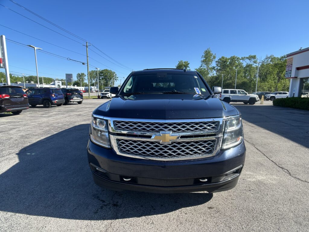 Front view of a dark blue Chevrolet pickup in a dealership lot with other cars and a clear blue sky behind it.