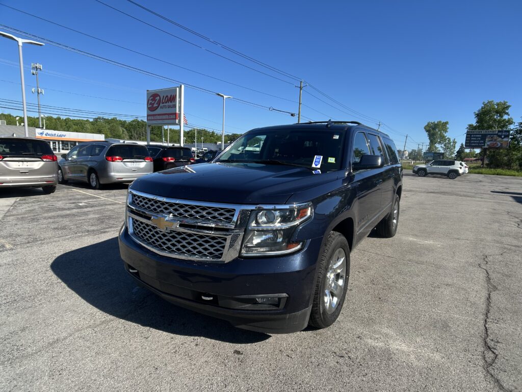 Front-right view of a dark blue SUV in a car dealership lot with several minivans parked nearby and an EZ Loan Auto Sales sign against a clear blue sky.
