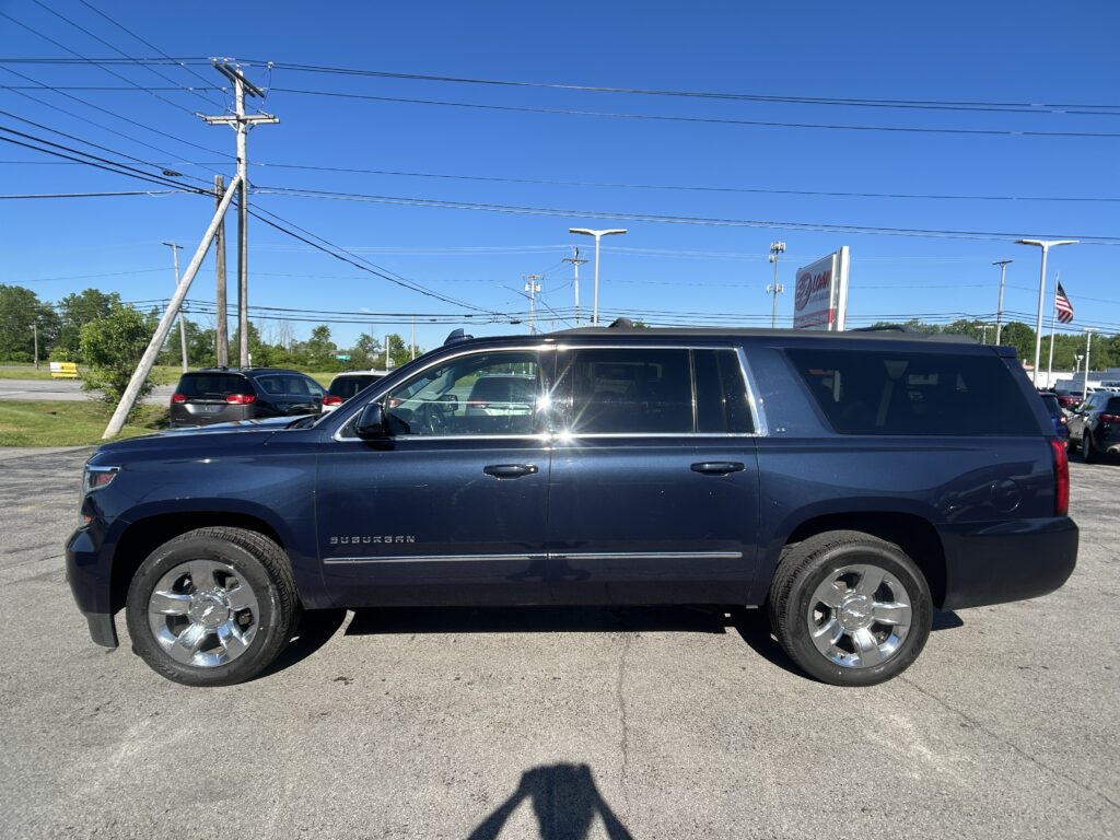 Dark blue SUV (Suburban) in a parking lot with power lines overhead and a clear blue sky behind it, dealership sign in the distance.