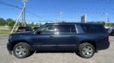 Dark blue SUV (Suburban) in a parking lot with power lines overhead and a clear blue sky behind it, dealership sign in the distance.