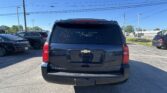 Rear view of a black Chevrolet Suburban in a parking lot, with other vehicles and power lines overhead on a sunny day, Suburban badge visible on the tailgate.