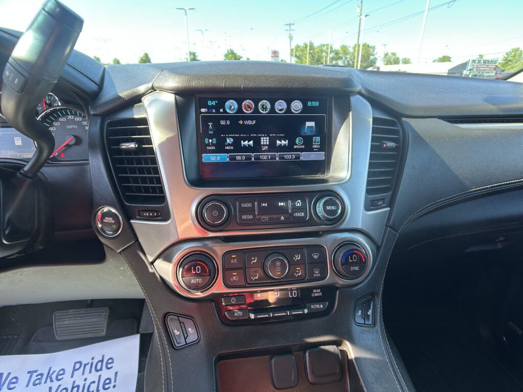 Inside view of a car dashboard showing a center touchscreen with radio controls and climate dials on the console below, and a steering wheel to the left.