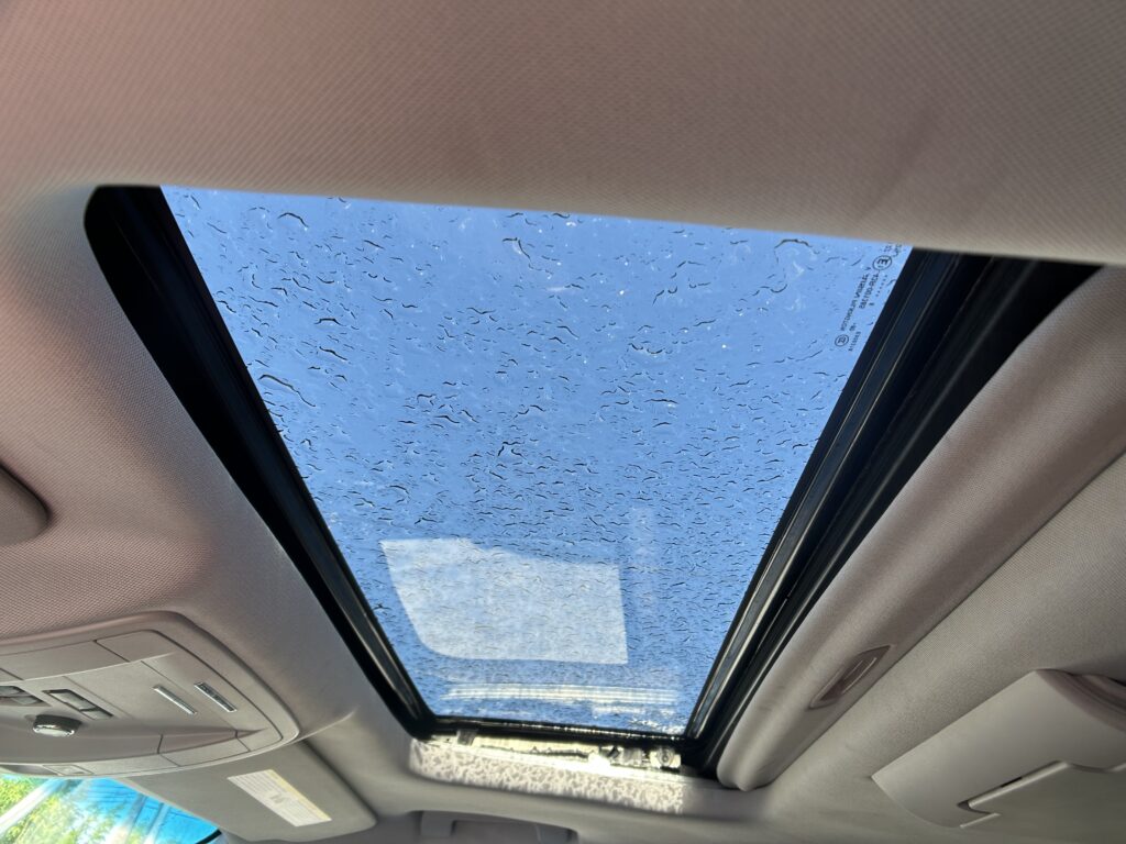 Inside a car looking up at an open panoramic sunroof with raindrops on the glass and blue sky beyond