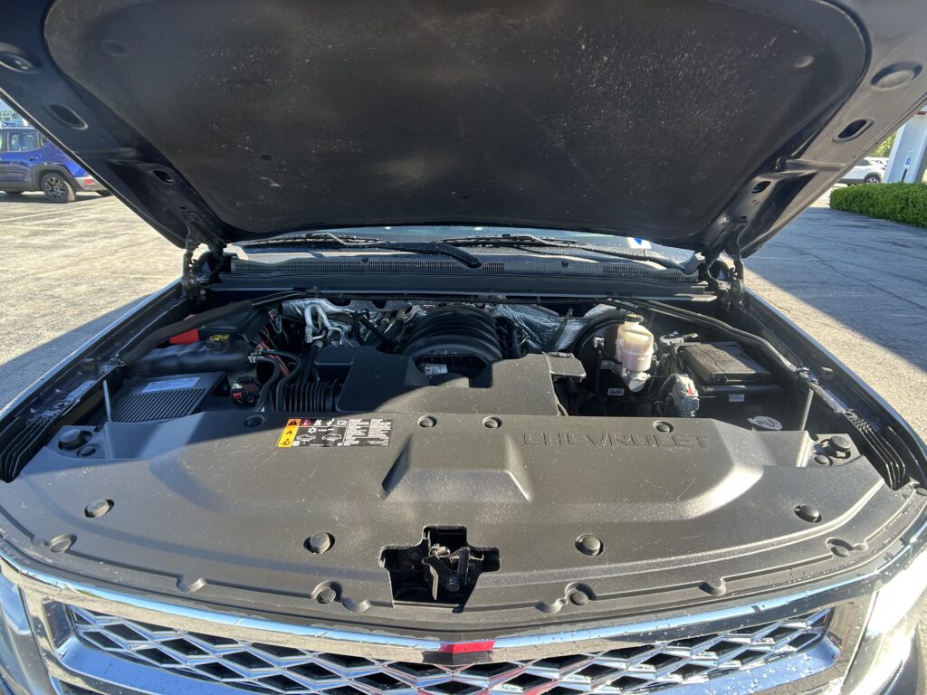 Open hood showing engine bay of a Chevrolet vehicle in a parking lot, with visible engine components and hoses.