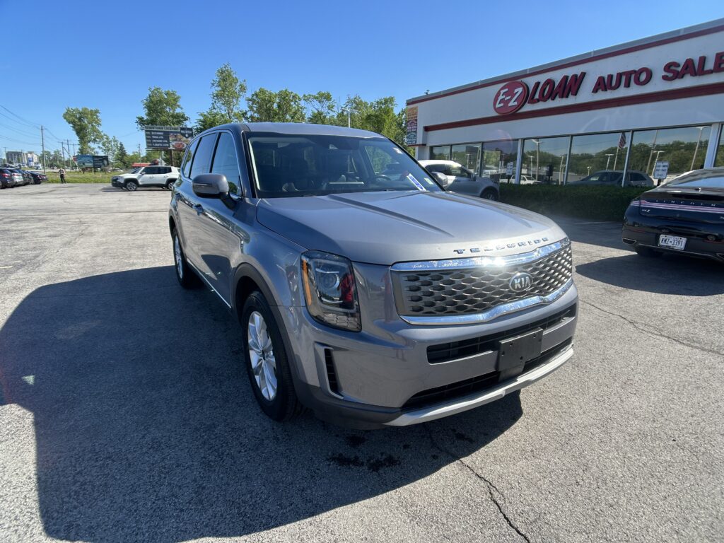 Front-right view of a silver Kia Telluride SUV parked in a car lot beside an auto dealership with bold EZ LOAN sign against a blue sky.