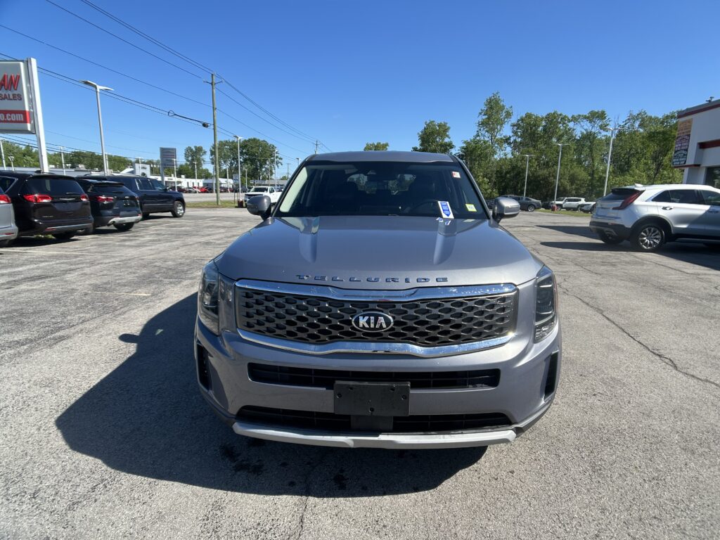 Front view of a silver Kia Telluride in a car dealership lot with other vehicles and a blue sky behind.