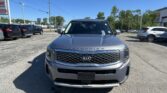 Front view of a silver Kia Telluride in a car dealership lot with other vehicles and a blue sky behind.